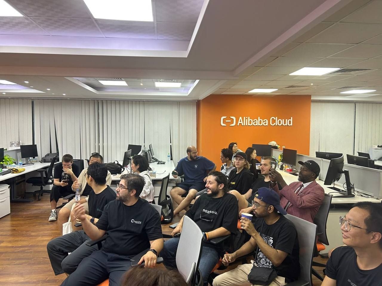 Group of people in a conference room with computers, orange Alibaba Cloud logo on the wall.