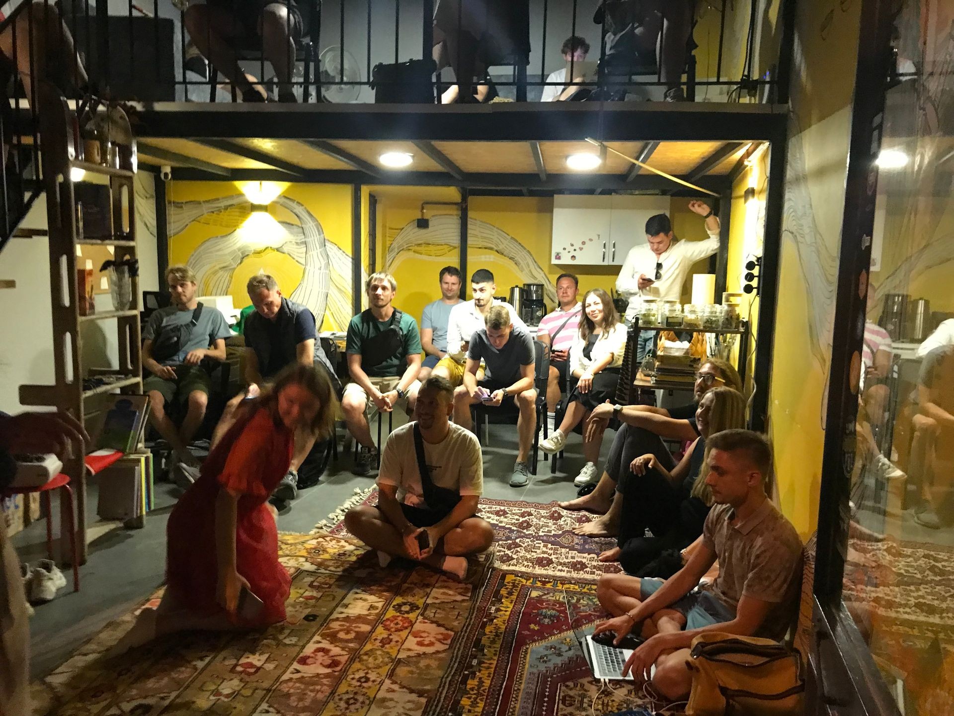 Group of people sitting and standing in a cozy room with yellow walls and a decorative carpet.