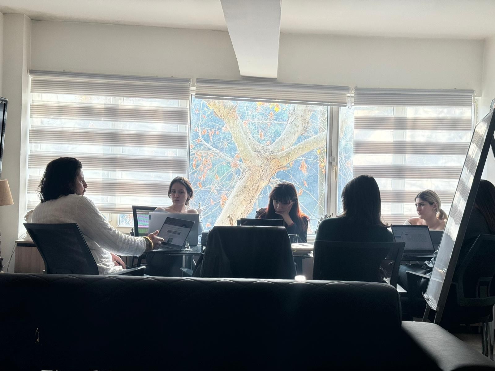 Group of people sitting at a table with laptops in a well-lit room with large windows.