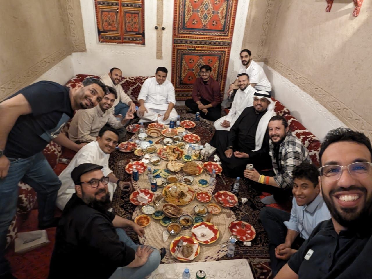 Group of people smiling around a table filled with dishes in a traditional room with patterned decor.