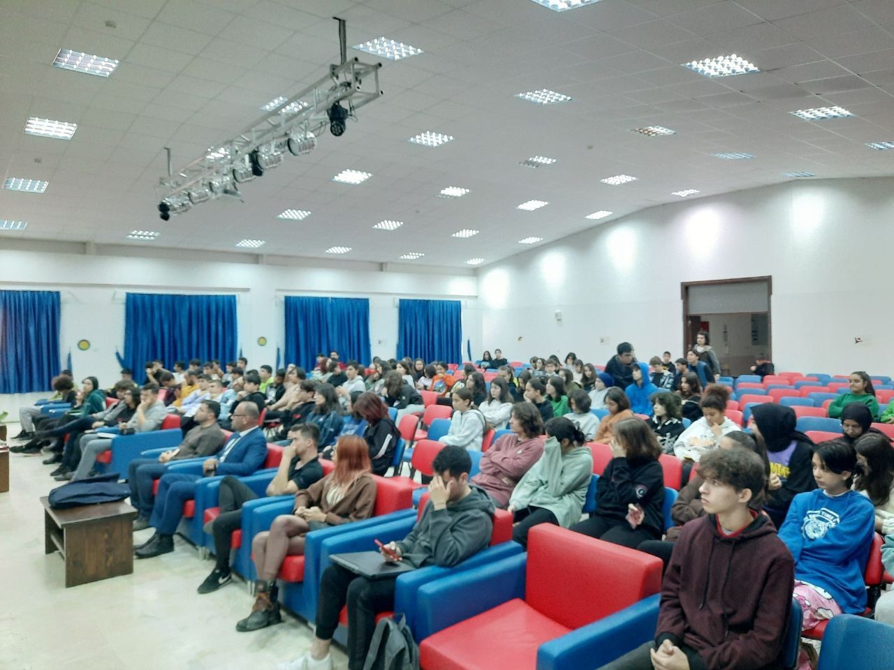 Audience seated in a colorful auditorium with blue curtains and bright lighting.