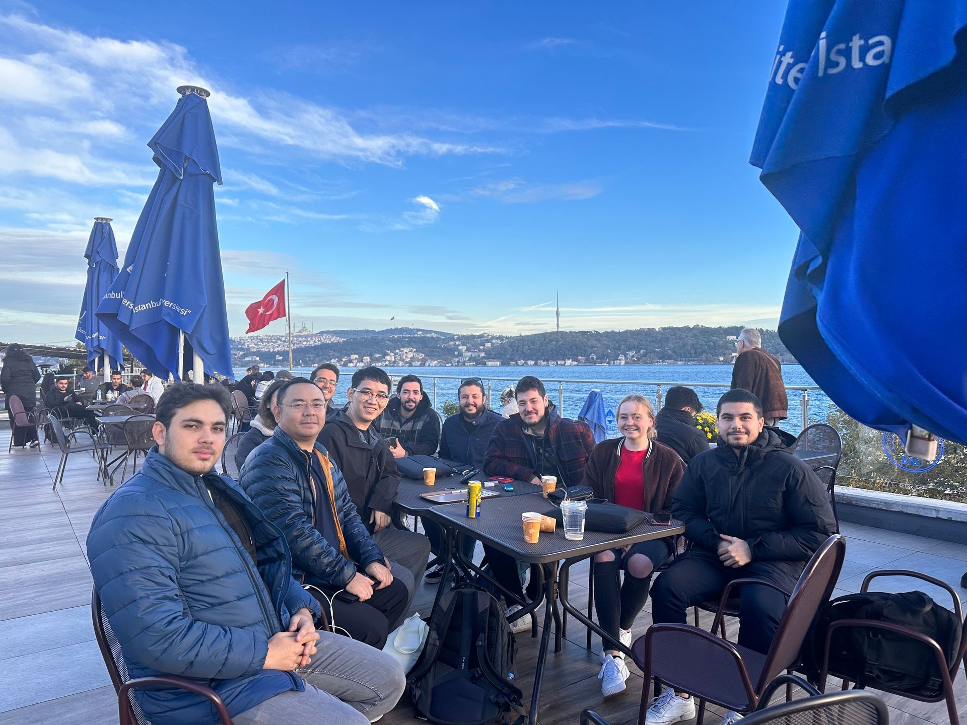 Group of people seated at an outdoor cafe by a waterfront with a Turkish flag in the background.