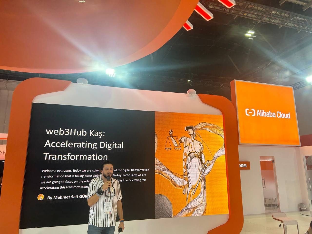 Person presenting at a digital transformation event with Alibaba Cloud signage and displays in the background.