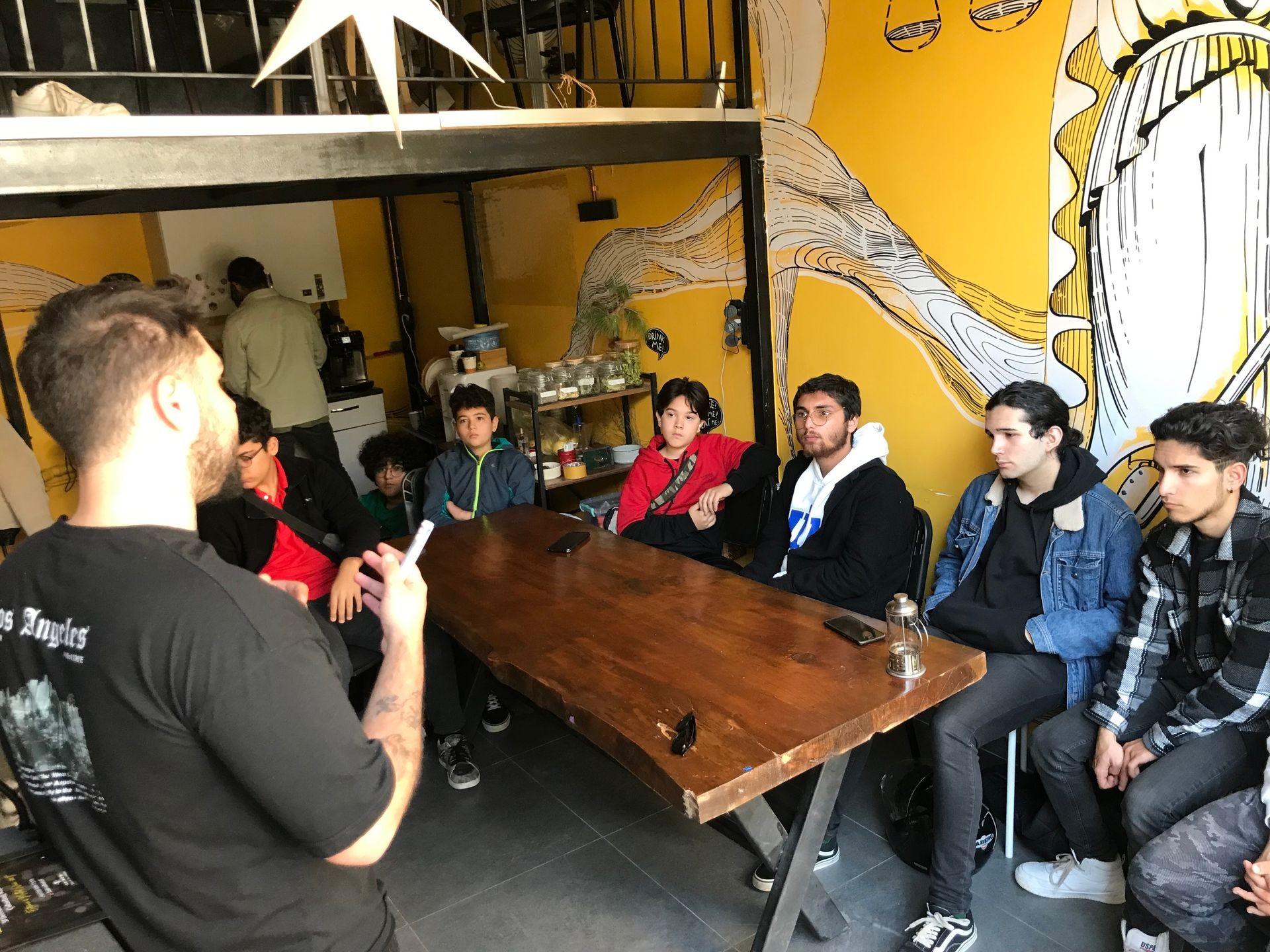 Person giving a talk to a group of attentive young people in a room with yellow mural walls.