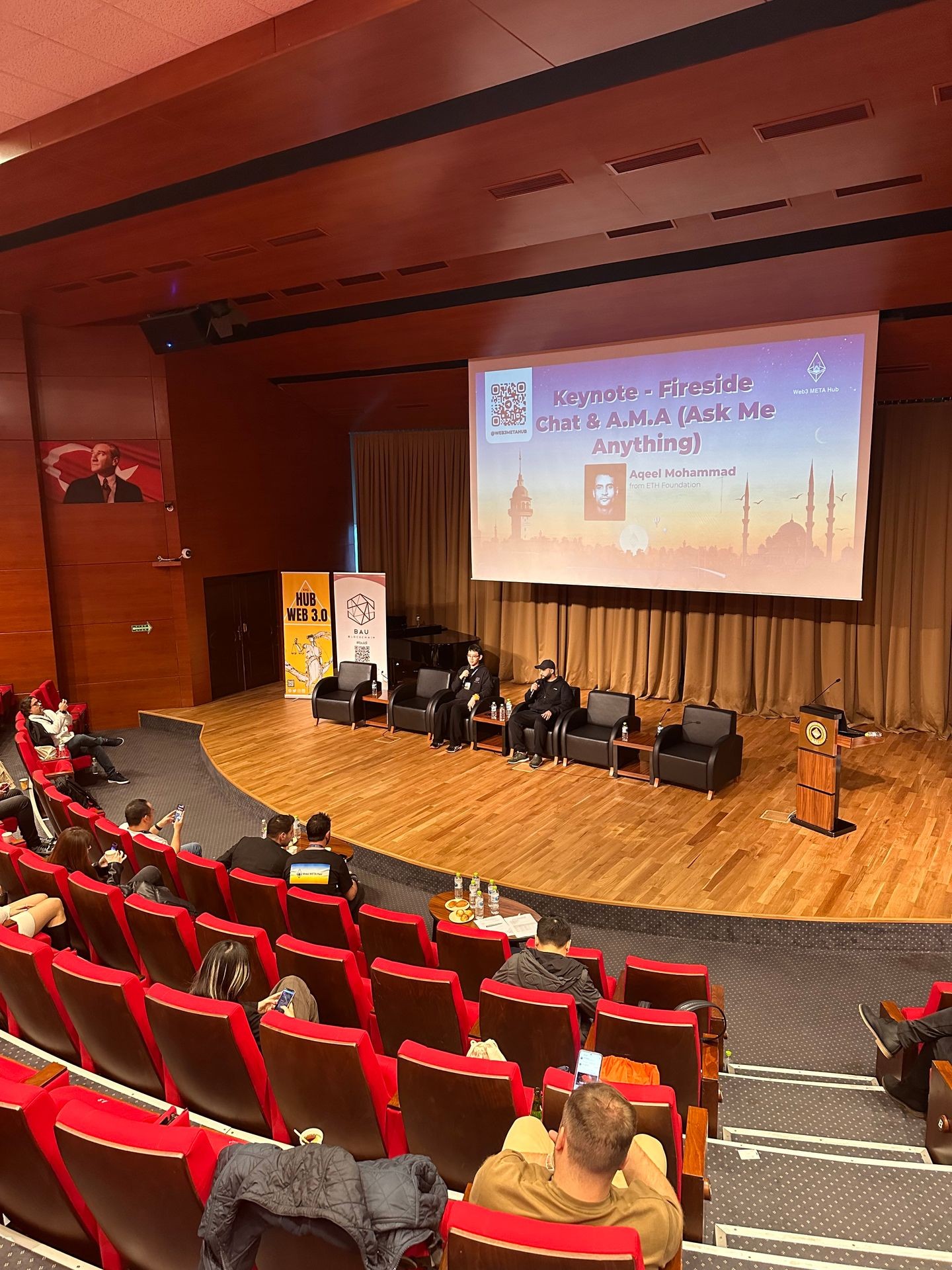 Conference hall with a keynote presentation on the screen and two speakers seated on stage.