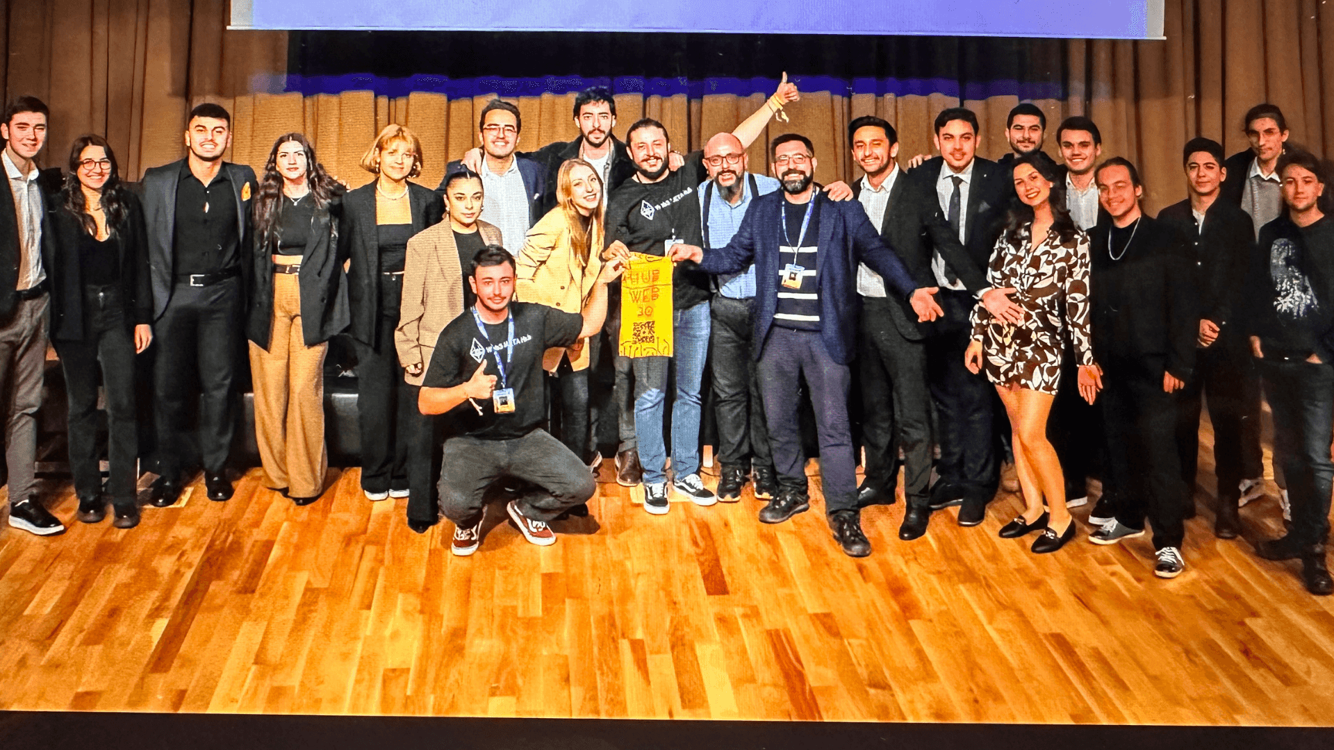 Group of people standing and posing together on a wooden stage with a large screen behind them.