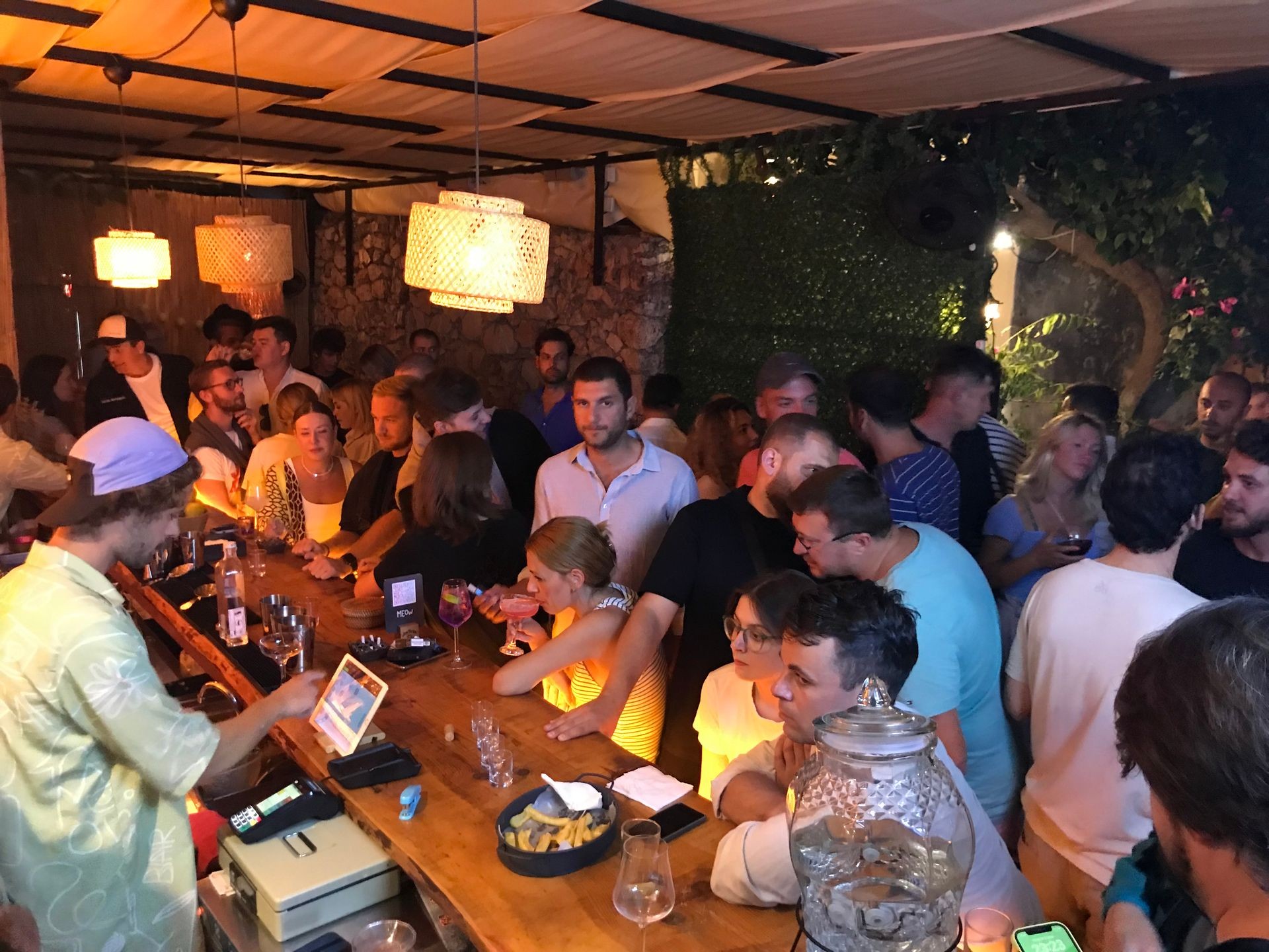 A crowded bar with people gathered around the wooden counter, illuminated by warm hanging lights.