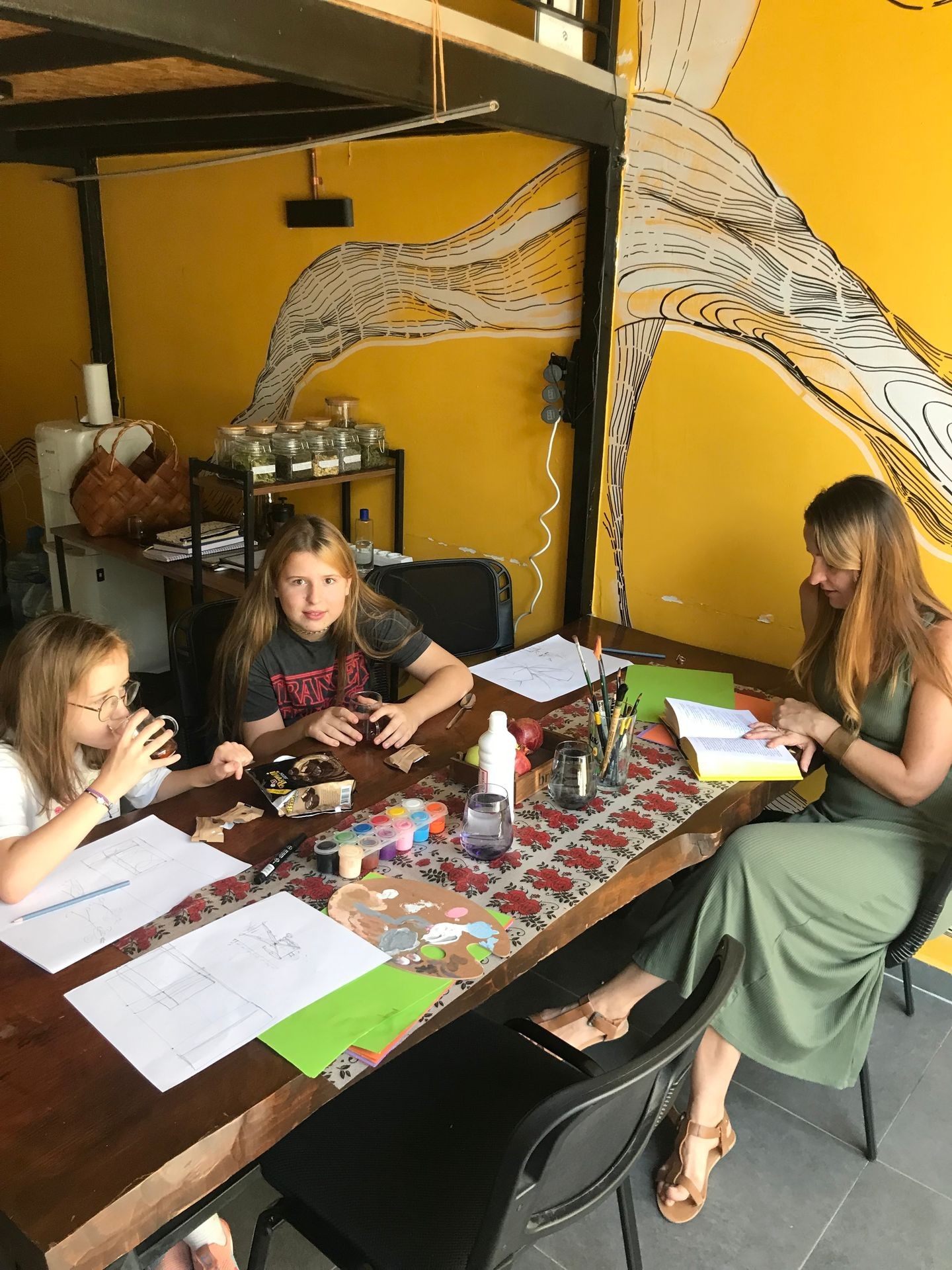 People sitting at a table with art supplies and sketches, against a yellow mural wall.