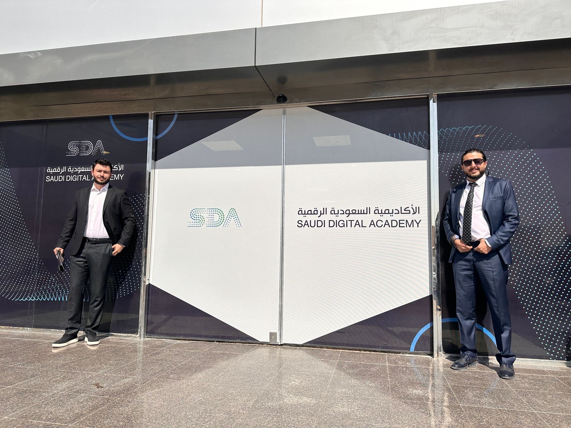 Two men in suits standing outside Saudi Digital Academy entrance.